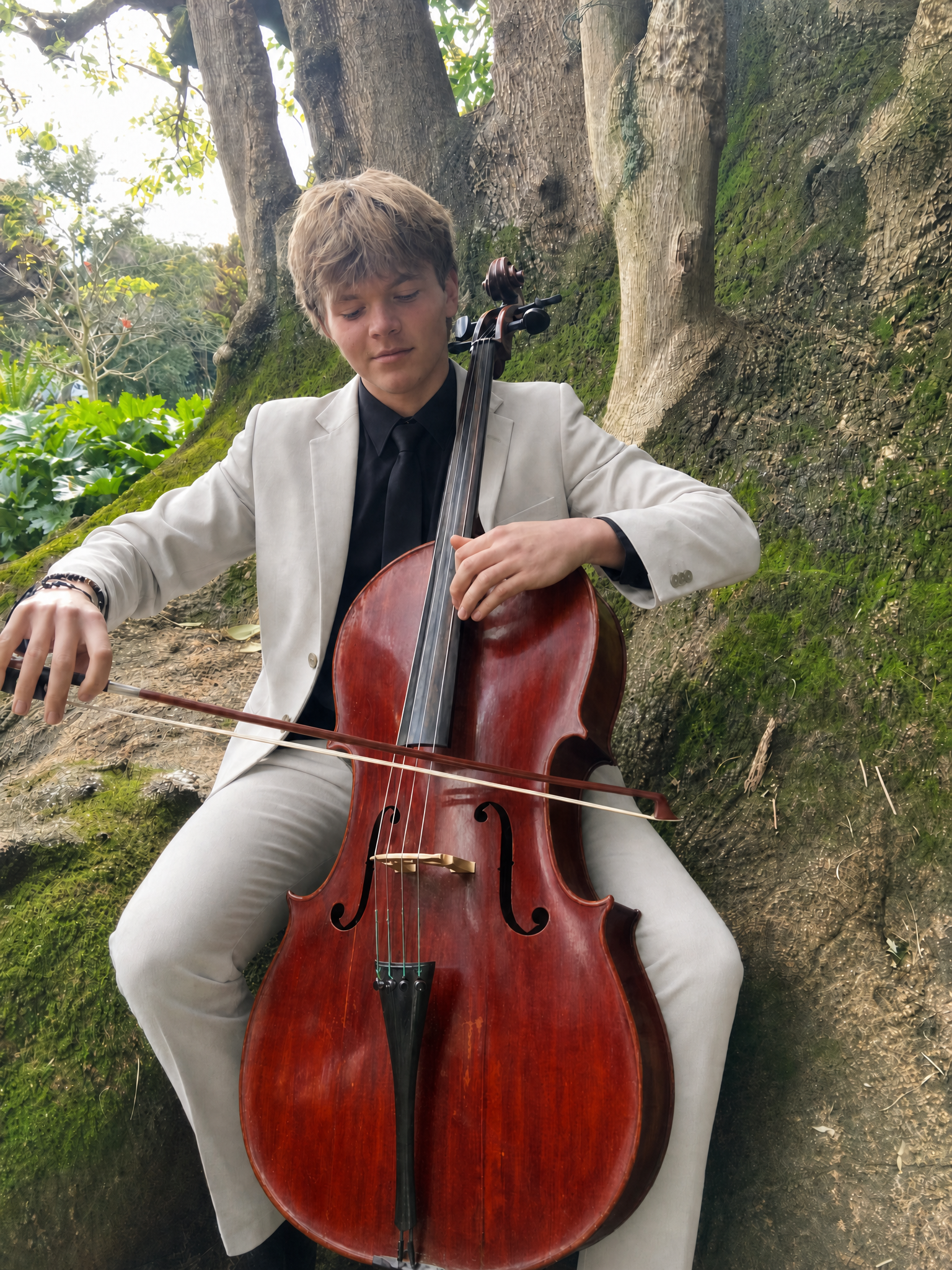 Luke Stamer playing cello beneath a large tree in a cream suit